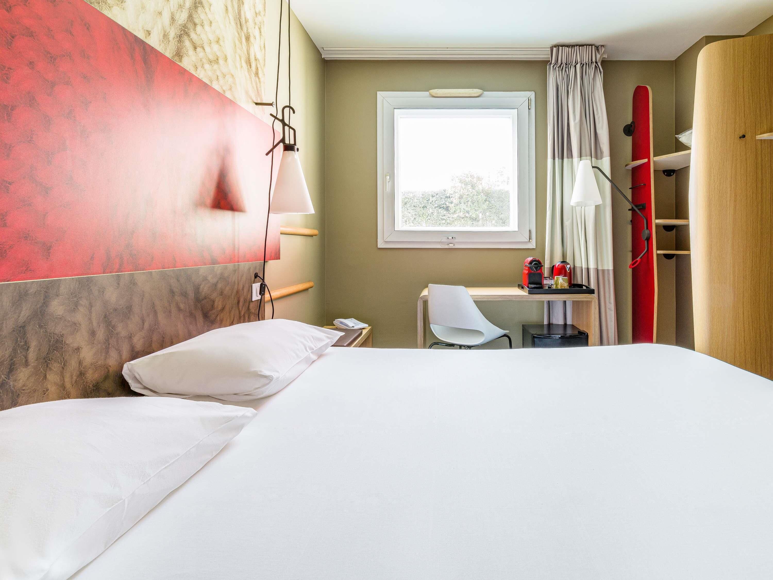 A hotel room with a bed, a desk, a chair, a window with curtains, and a surfboard.