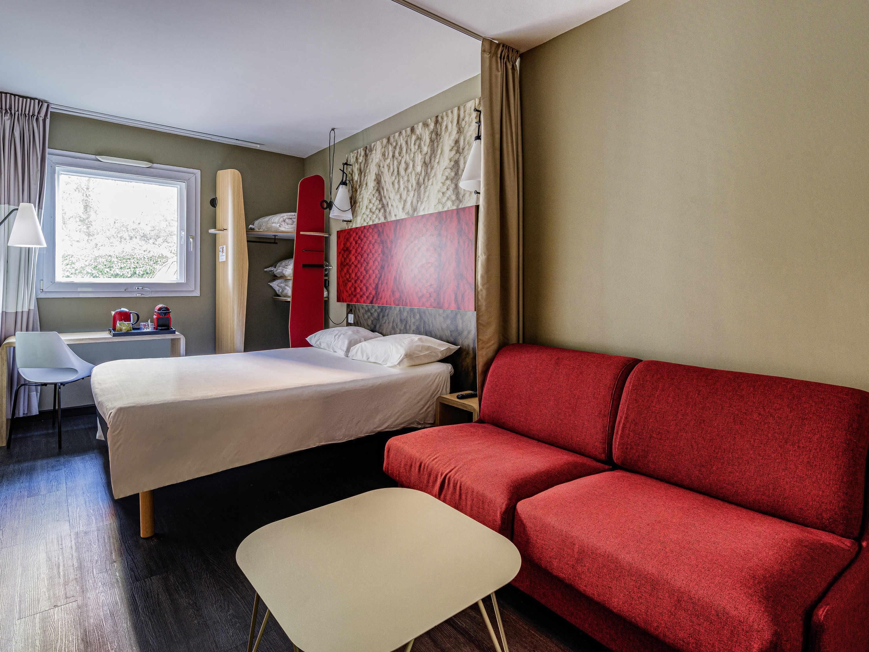 A hotel room with a bed, a red sofa, a small table, a chair, a desk with a lamp, and a window.