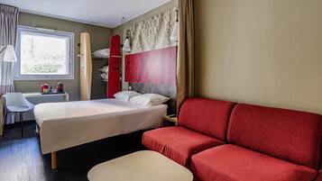 A hotel room with a bed, a red sofa, a small table, a chair, a desk with a lamp, and a window.