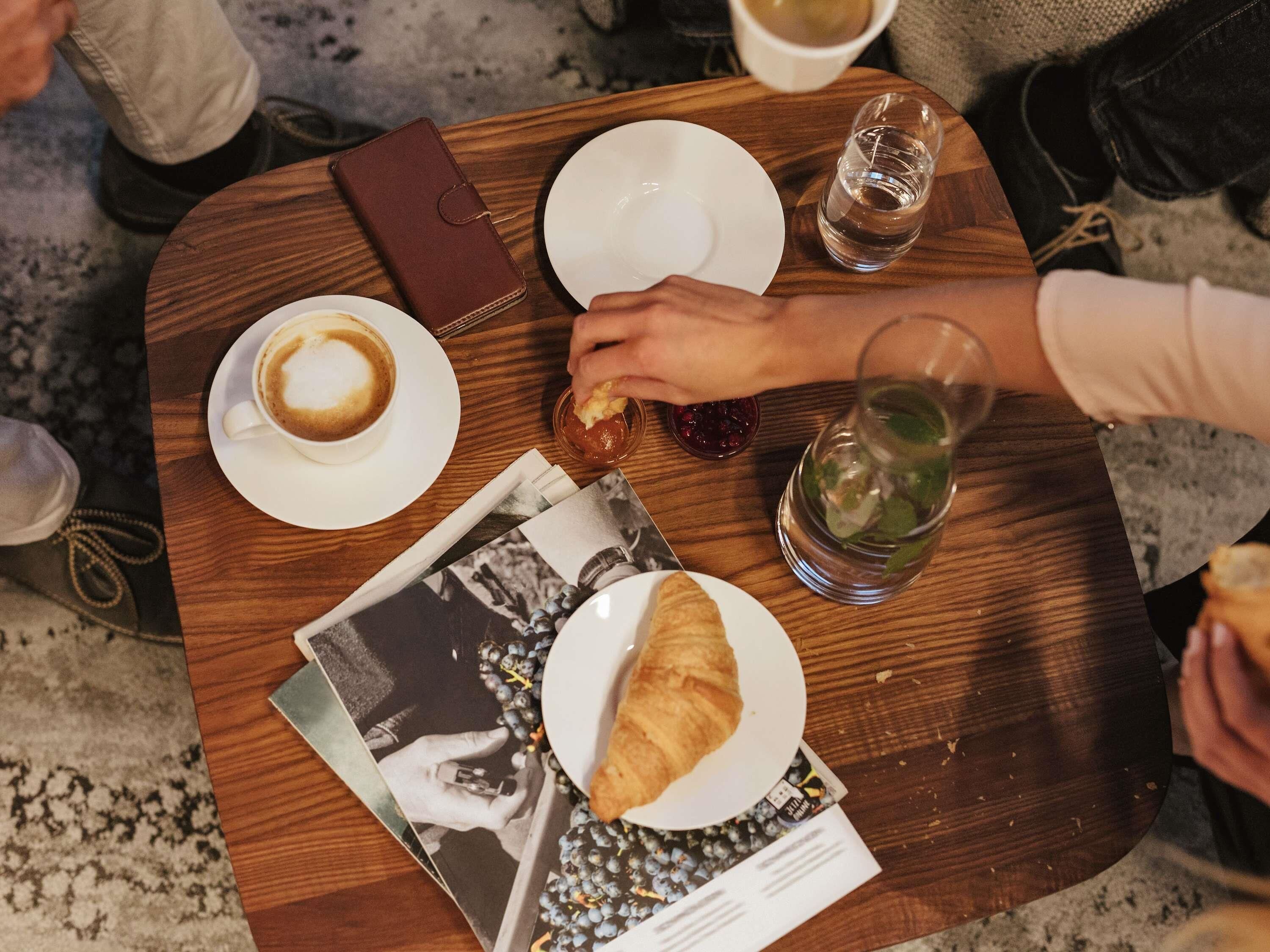 A wooden table with a croissant, a cup of coffee, a glass of water, and a magazine.