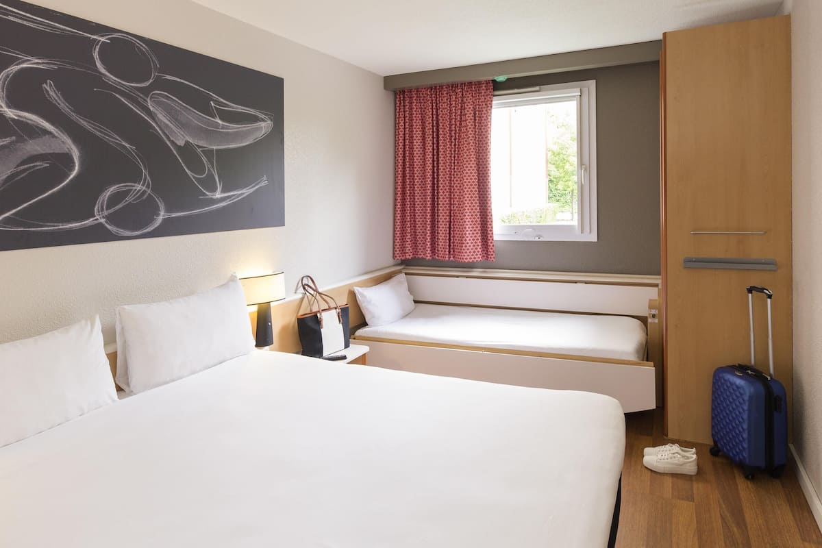 Ein Hotelzimmer mit einem großen Bett, einem kleinen Sofa, einem Fenster mit roten Vorhängen, einem Wandbild und einem Koffer.