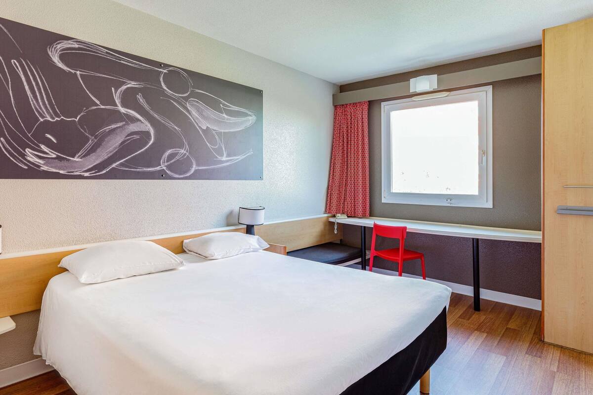 Ein Hotelzimmer mit Bett, Schreibtisch, Stuhl, einem Fenster mit Vorhängen und einem Wandbild.