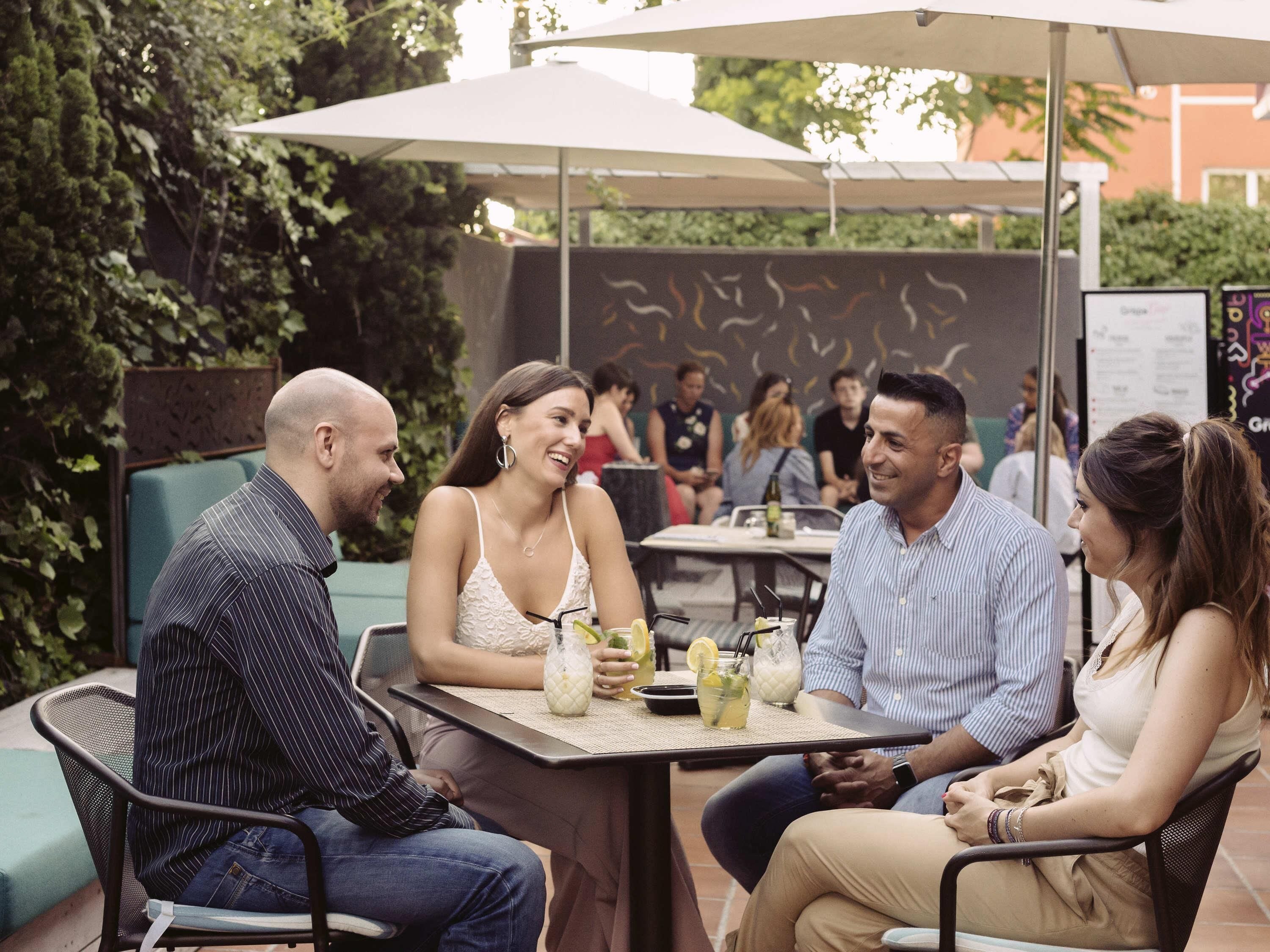 Un grupo de personas sentadas en una mesa al aire libre, con bebidas, rodeadas de follaje y sombrillas.