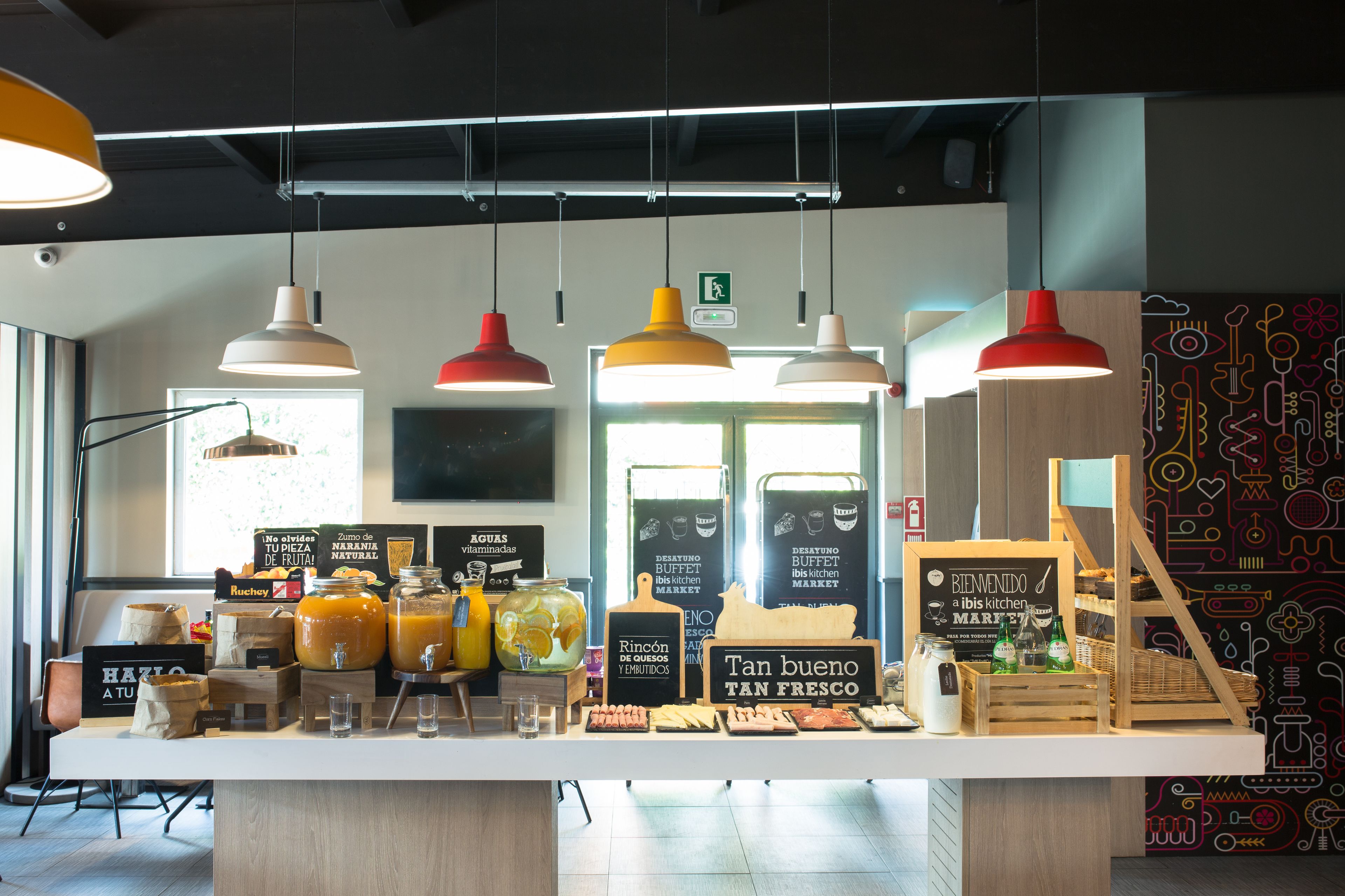A cafe counter with various food items and drinks, hanging pendant lights, and a wall with a colorful abstract design.