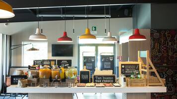 Una barra de café con distintos alimentos y bebidas, lámparas colgantes y una pared con un diseño abstracto y colorido.