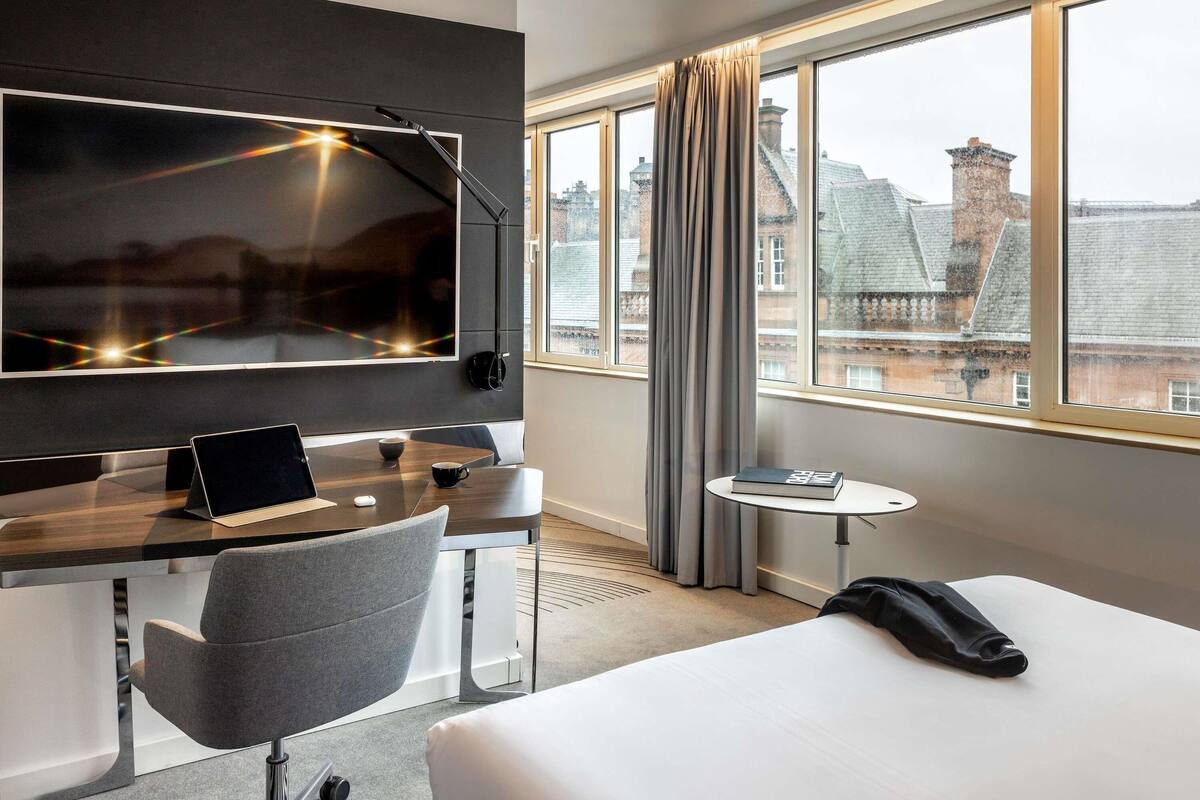 A hotel room with a bed, a desk with a laptop, a chair, a small table with a book, a TV mounted on the wall, and a window with curtains.