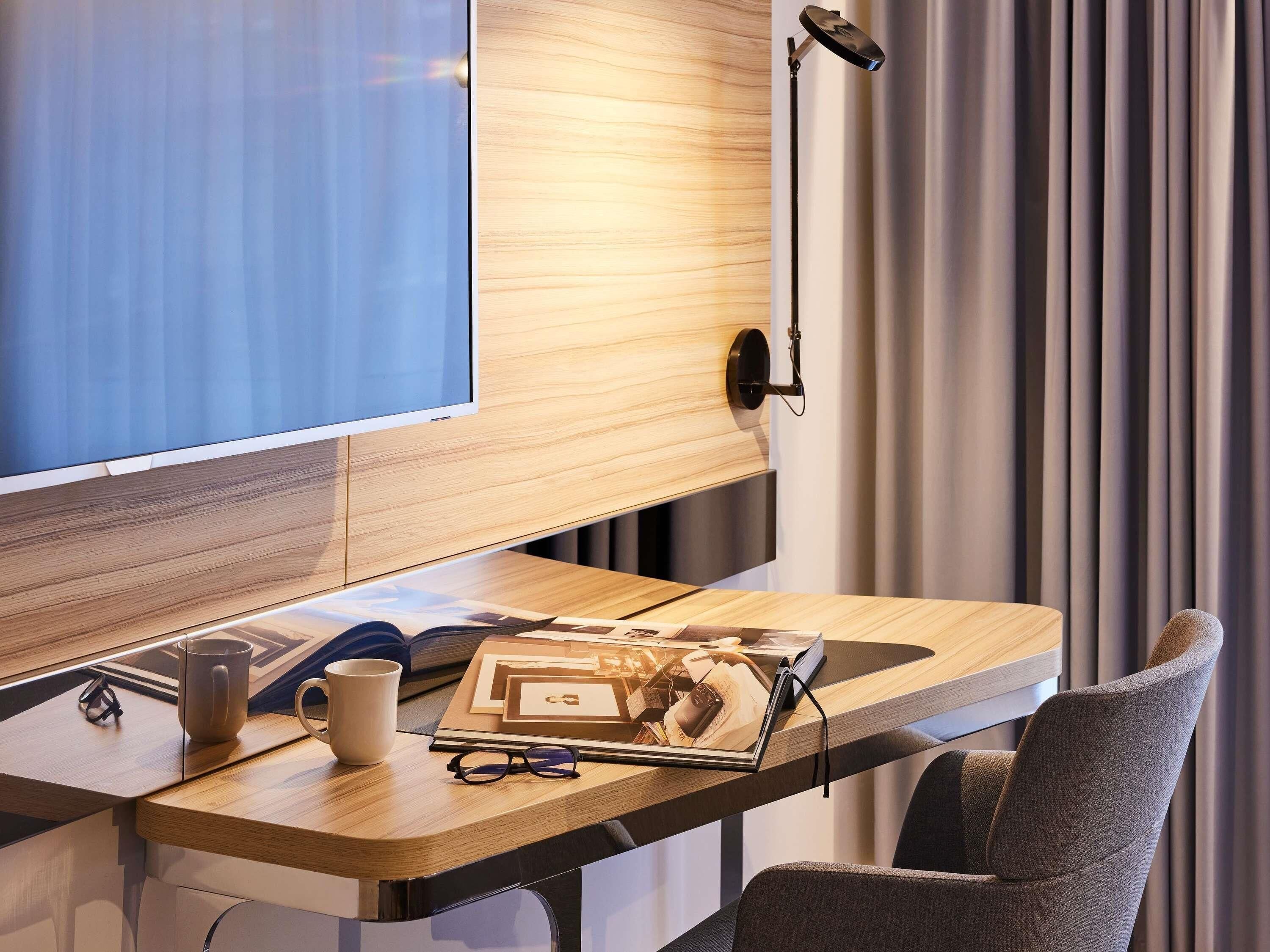 A modern hotel room with a wooden desk, a flat-screen TV, a lamp, and a comfortable chair.