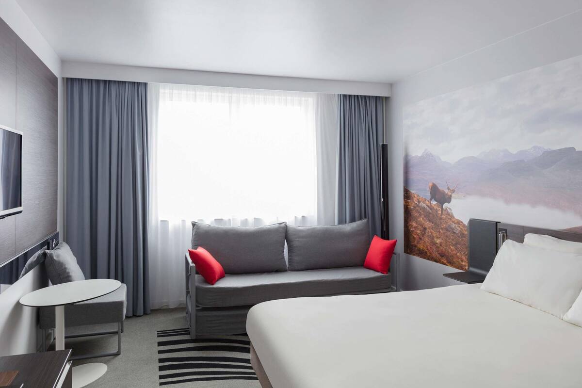 A modern hotel room with a large bed, a sofa bed, a desk, and a TV.