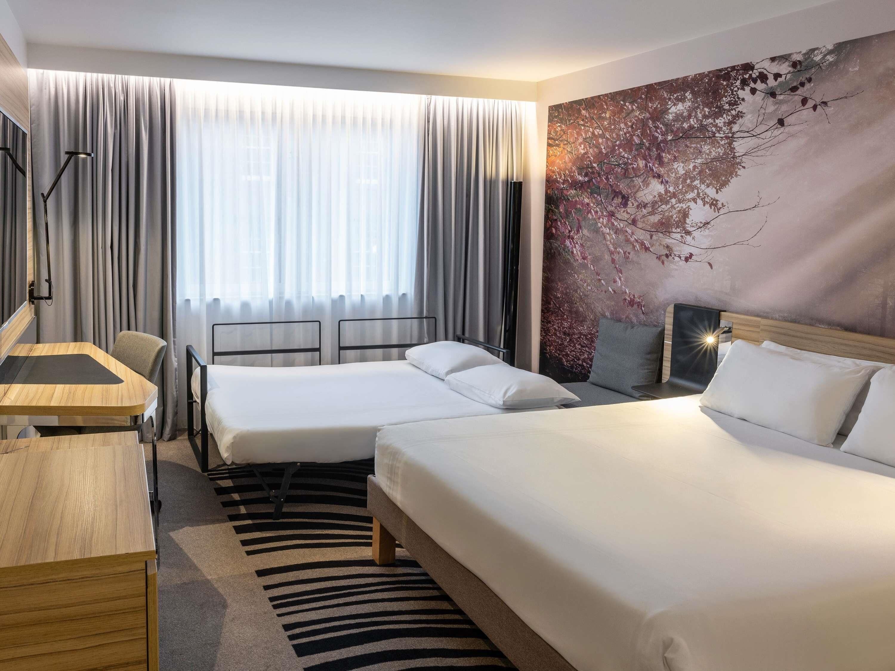 A hotel room with two beds, a desk, a chair, and a large window with curtains.