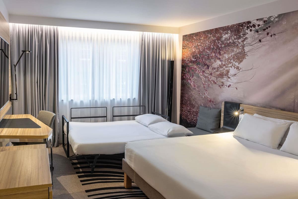 A hotel room with two beds, a desk, a chair, and a large window with curtains.