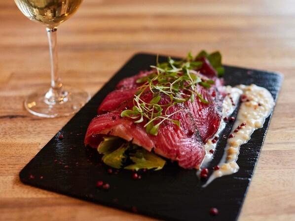 A black slate plate with a steak, greens, and sauce, accompanied by a glass of white wine on a wooden table.