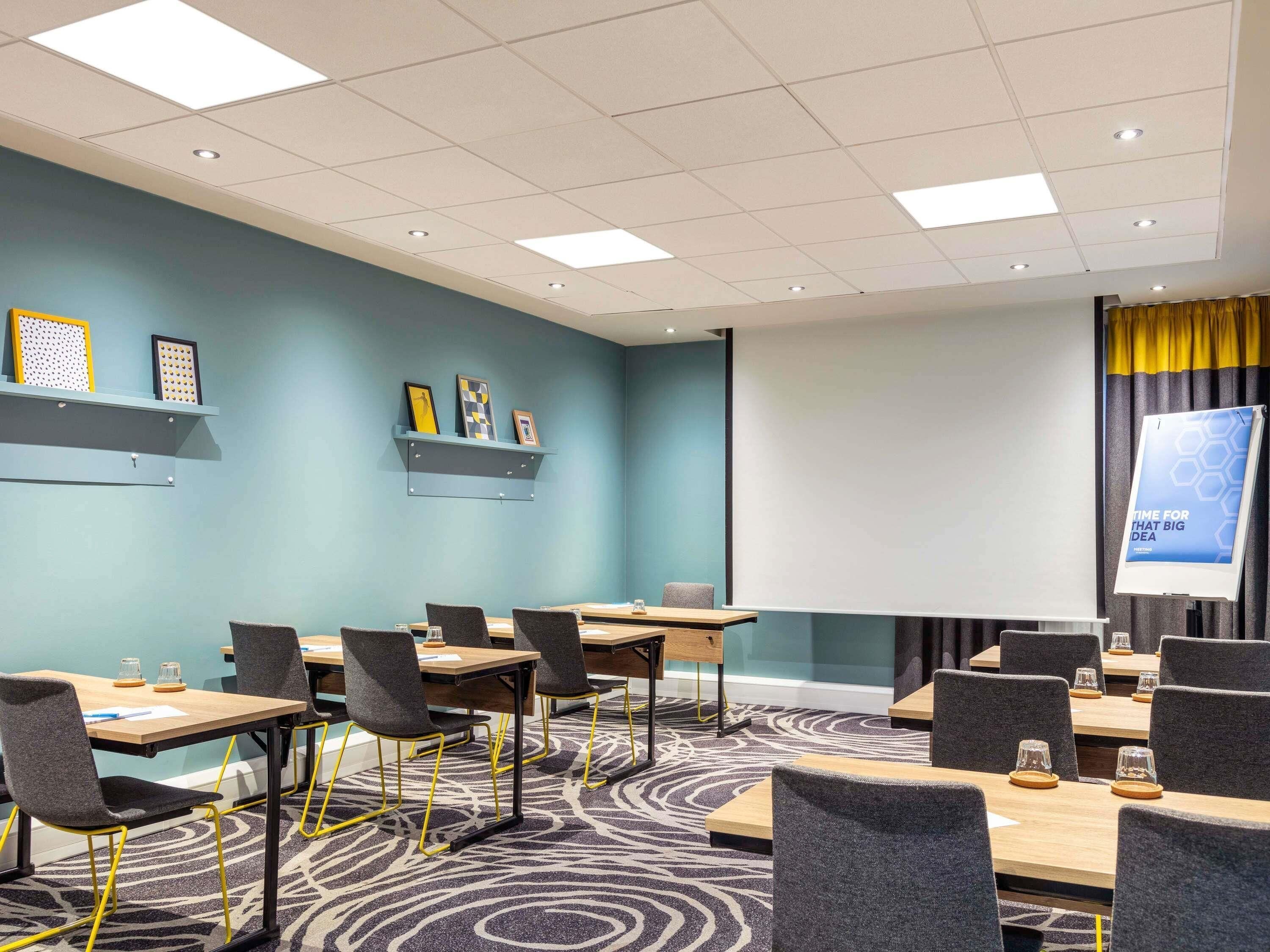 Een vergaderzaal met tafels, stoelen, een projectiescherm en een vloerbedekking.