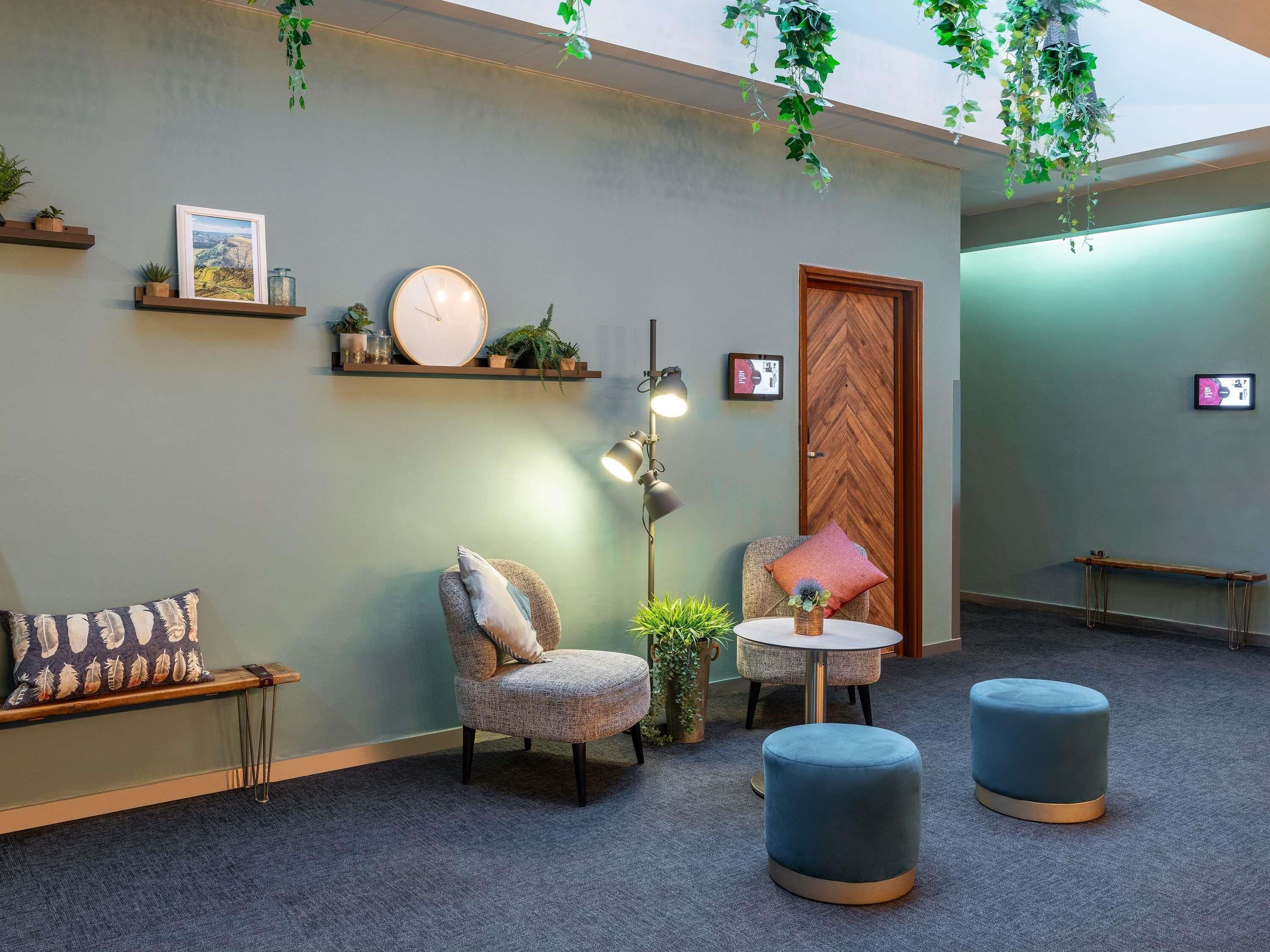 A modern room with a sofa, armchair, ottoman, and a round table. There are shelves with plants and a clock on the wall.