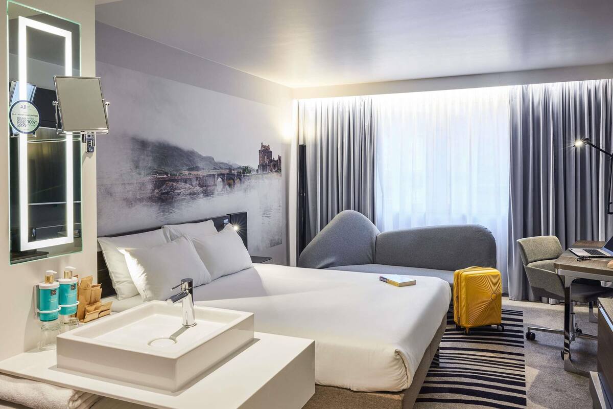 A hotel room with a bed, a desk, a chair, a bathroom with a sink, and a large window with curtains.