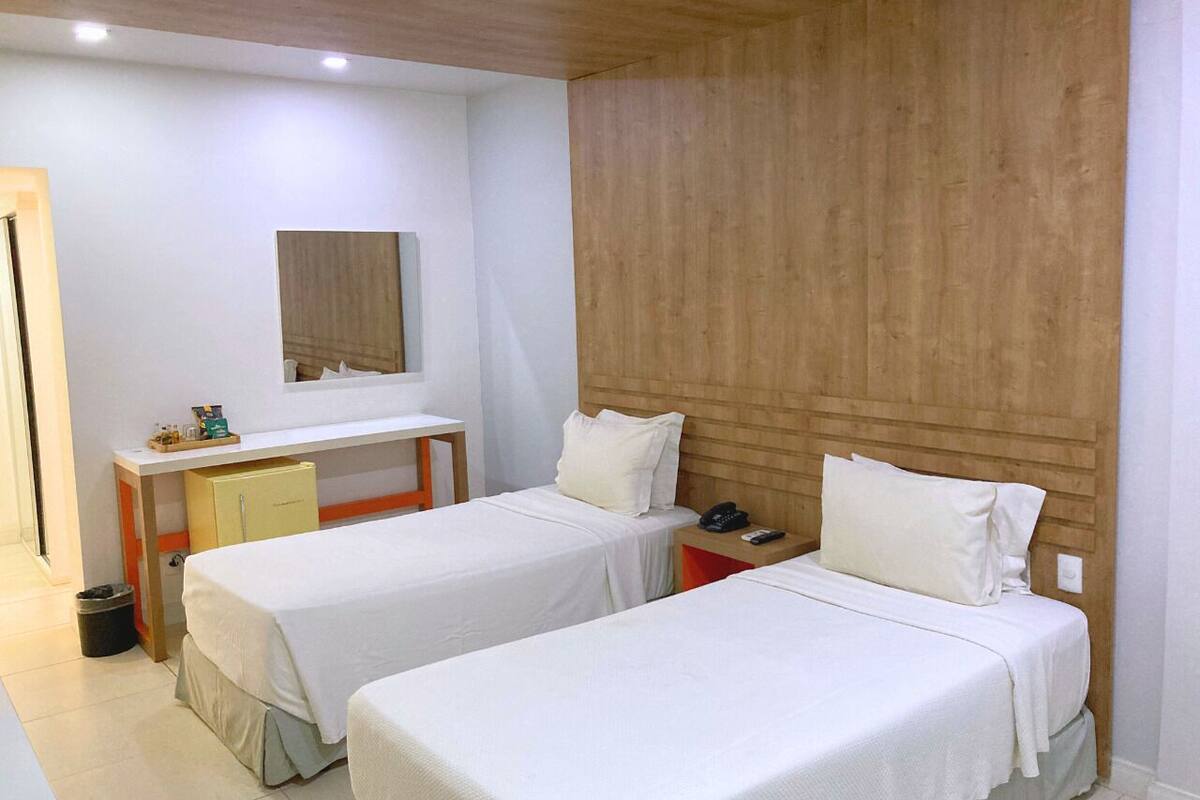 Une chambre d’hôtel avec deux lits simples, un bureau avec une chaise, un petit placard et un miroir au mur.