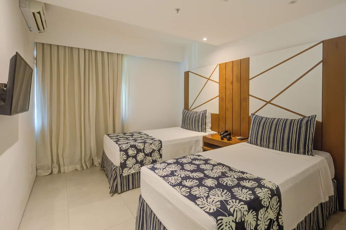 Une chambre d’hôtel avec deux lits, une télévision, une tête de lit en bois et une grande fenêtre avec des rideaux.