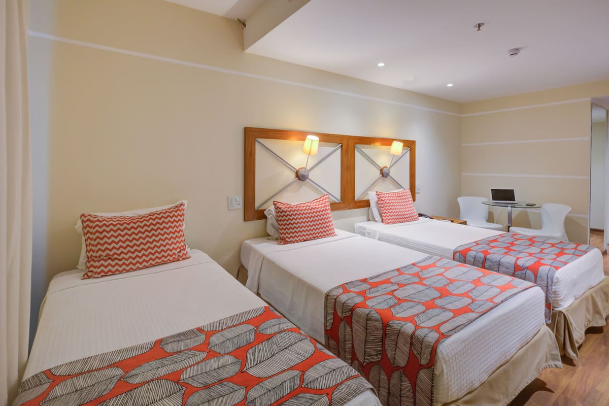 Une chambre d’hôtel avec trois lits simples, chacun agrémenté d’une couvre-lit à motifs et de coussins assortis. On y trouve un petit bureau avec un ordinateur portable, une chaise et un miroir au-dessus du lit.