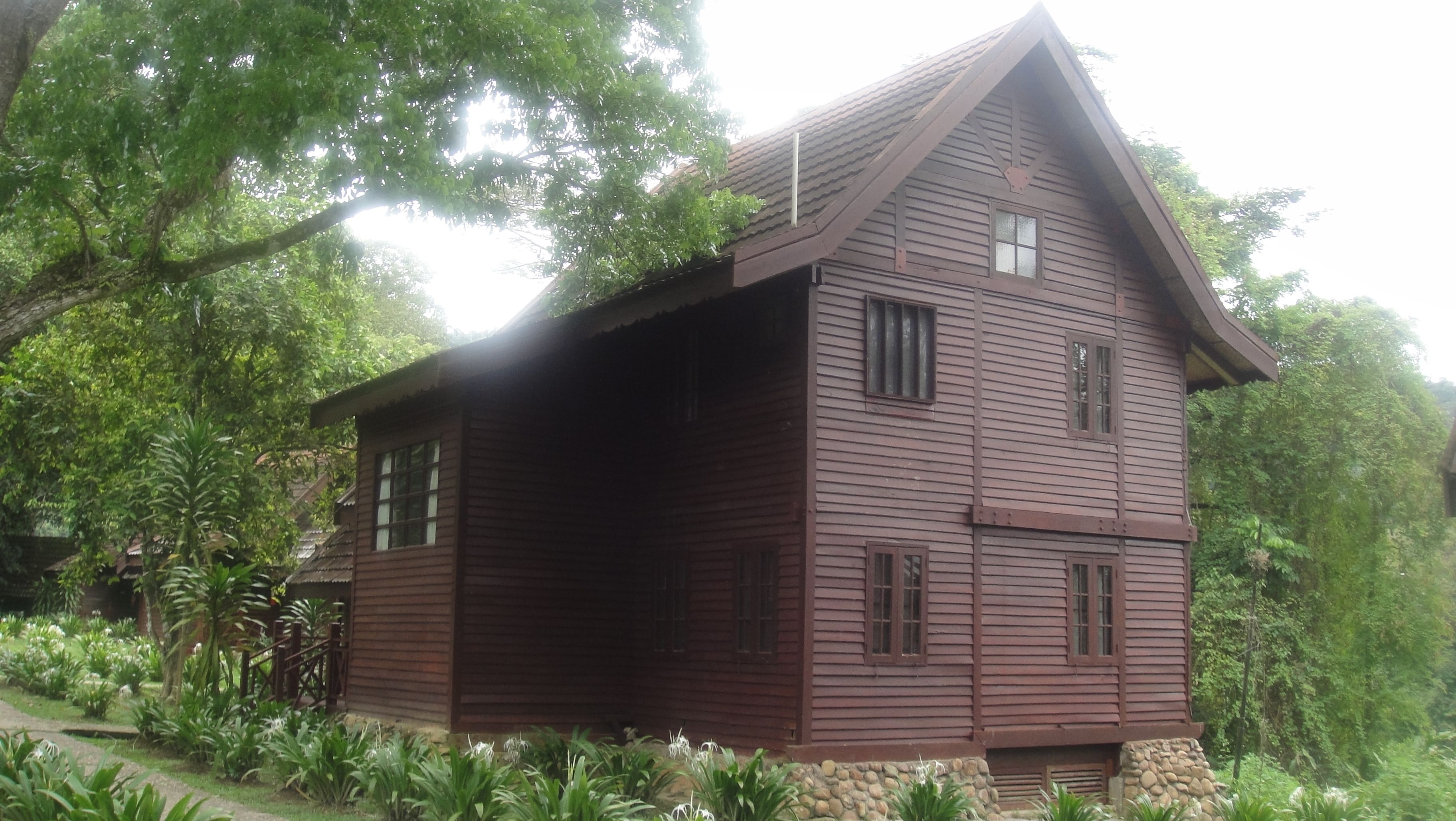 A two-story wooden cabin with a steep gabled roof, surrounded by lush greenery.