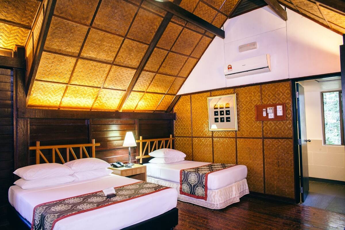 Een kamer met twee bedden, een houten plafond, een airconditioningapparaat, een nachtkastje met een lamp, en een elektrisch paneel aan de muur.