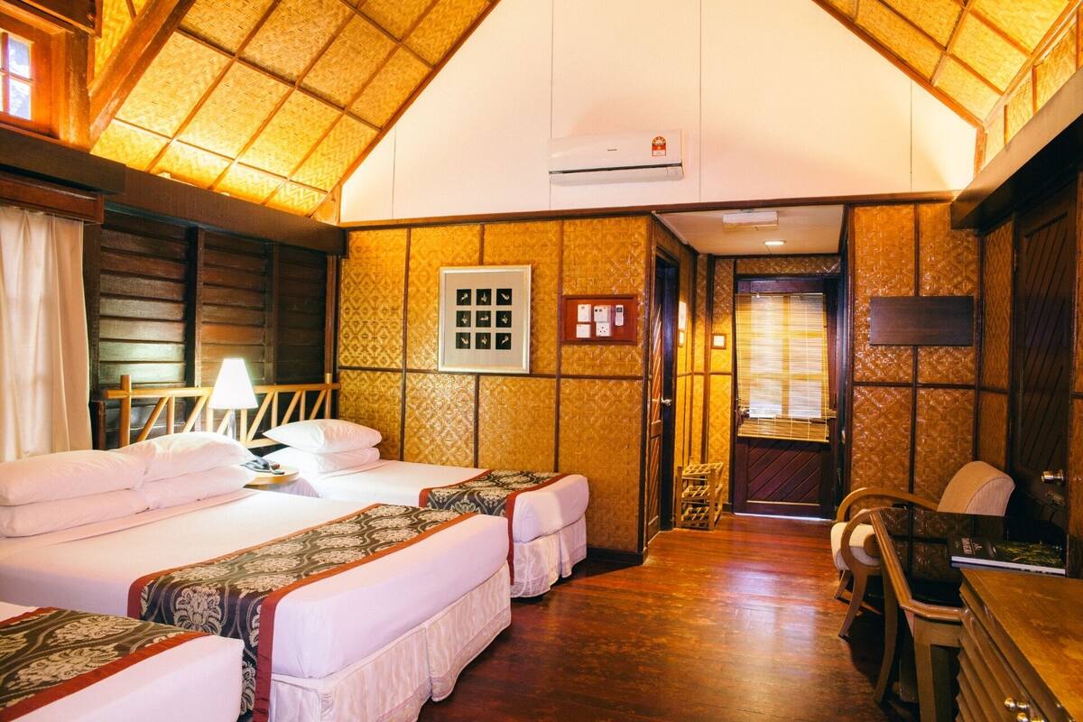 Een hotelkamer met een hoge plafond, houten balken en een groot bed met een geborduurd beddegoed. Er is een bureau met een stoel, een raam met jaloezieën, en een lijst met een foto aan de muur.