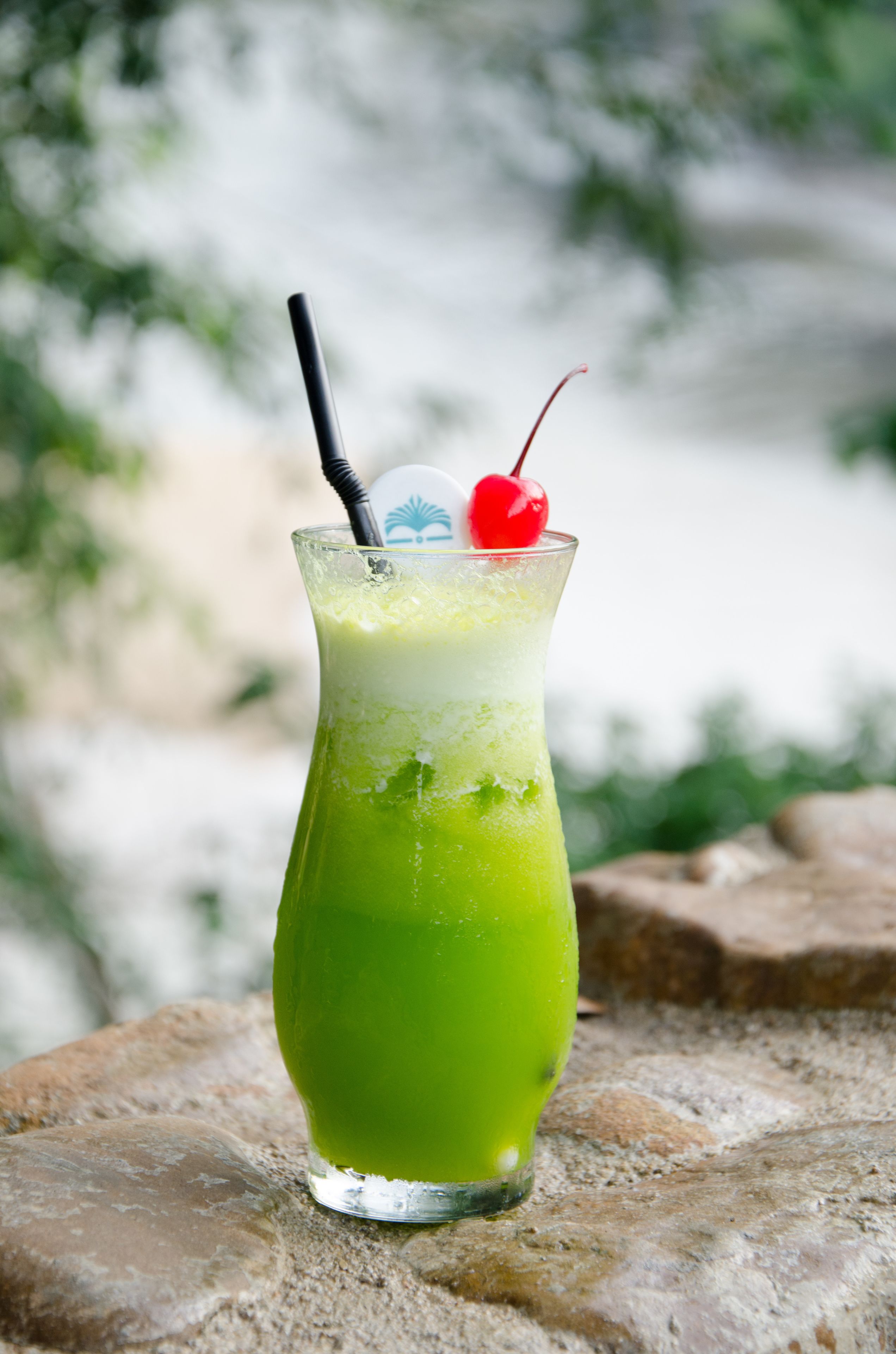 A tall glass with a green beverage, a straw, and a cherry on top, placed on a stone surface.