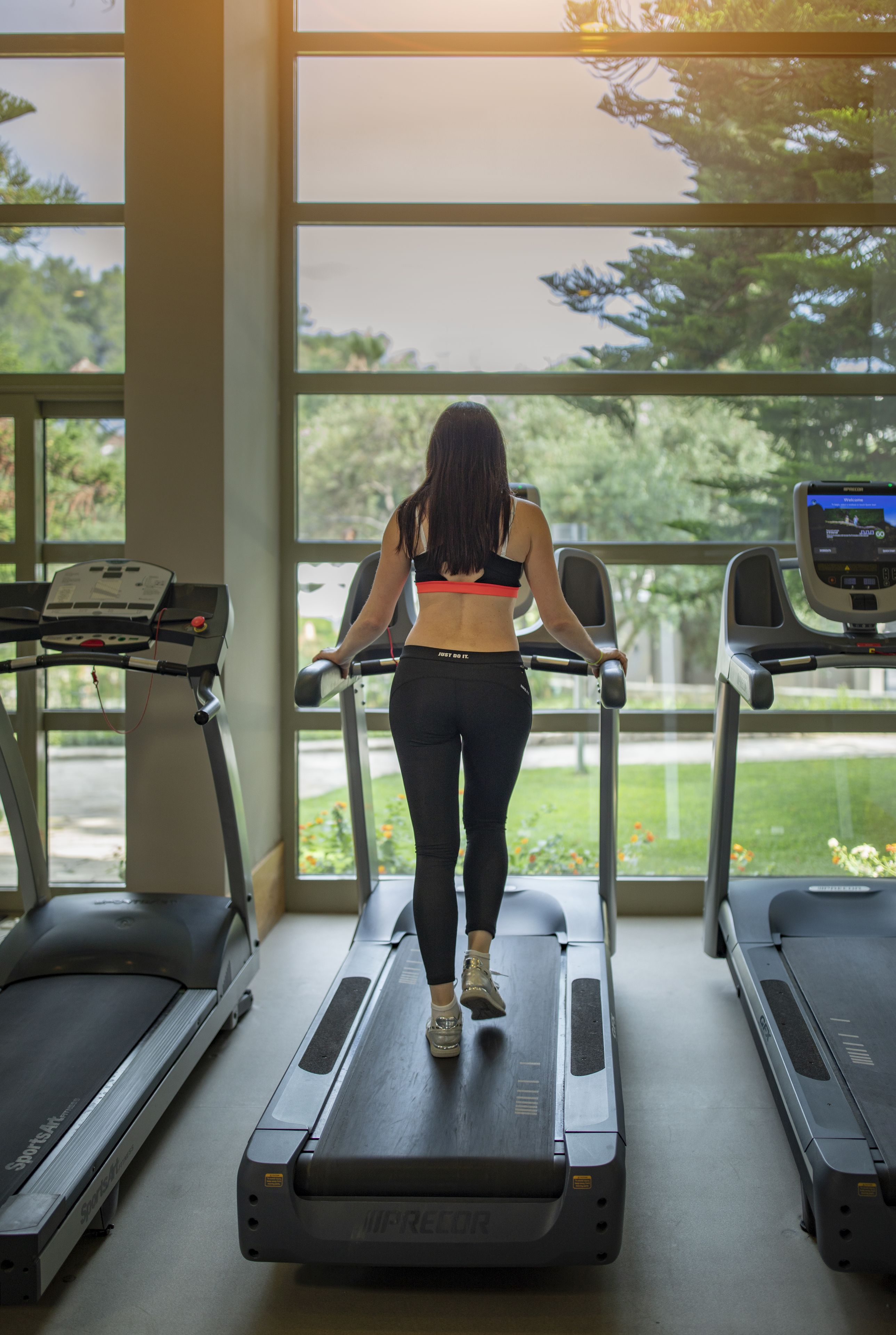Eine Frau läuft auf einem Laufband in einem Fitnessstudio mit großen Fenstern. Im Hintergrund ist ein weiteres Laufband zu sehen.