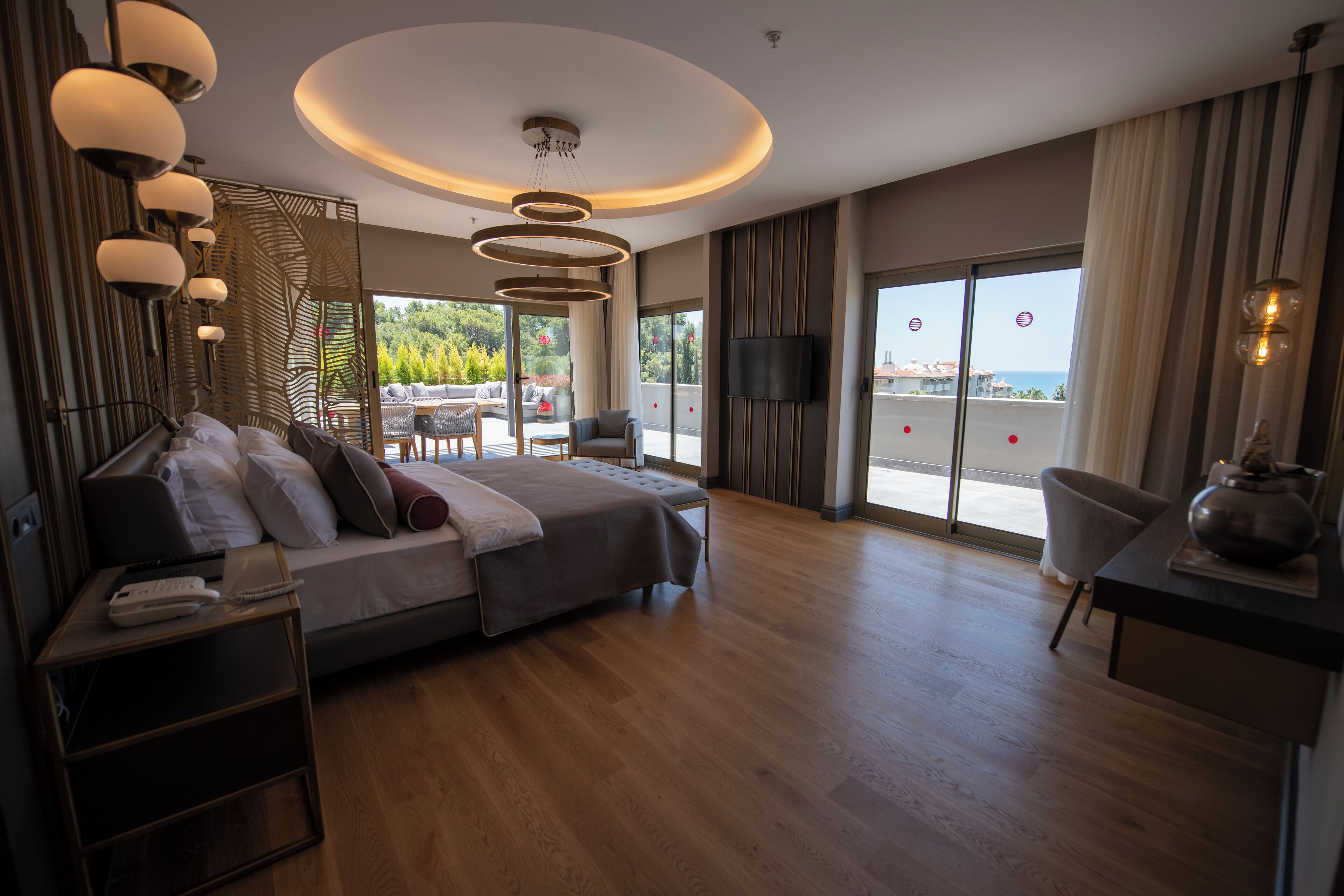 A modern hotel room with a large bed, a desk, and a TV. There is a balcony with a view of the sea.