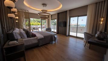 A modern hotel room with a large bed, a desk, and a TV. There is a balcony with a view of the sea.