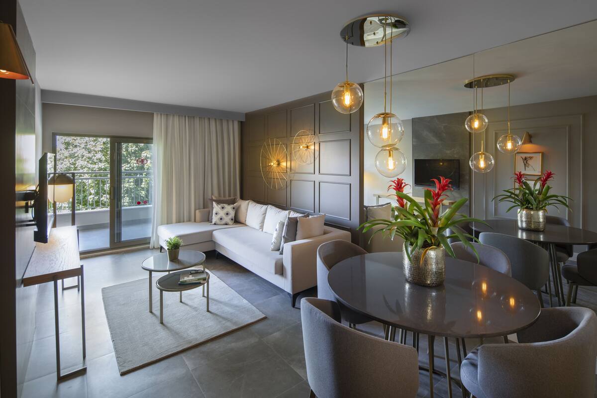 A modern living room with a sofa, dining table, and chairs. There are potted plants and a television on the wall.