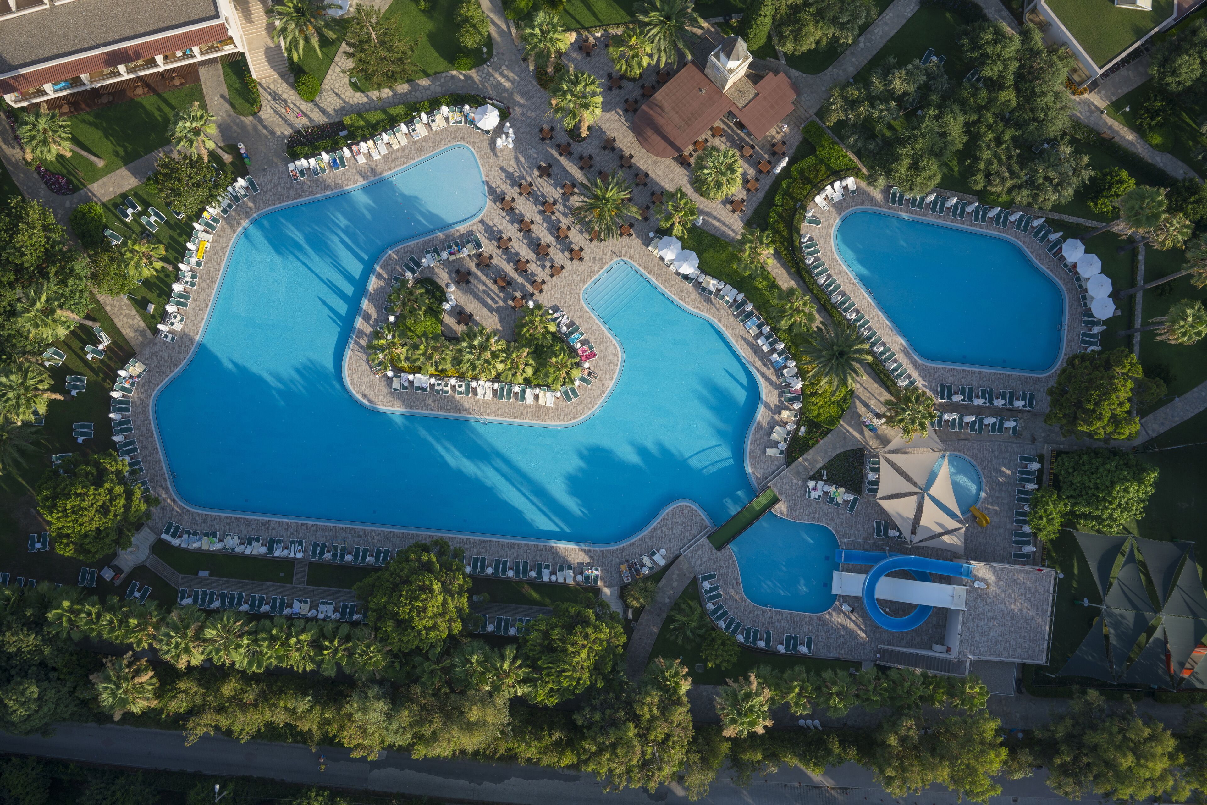 An aerial view of a resort with two large swimming pools, surrounded by lounge areas and palm trees.