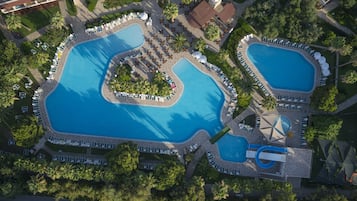 An aerial view of a resort with two large swimming pools, surrounded by lounge areas and palm trees.