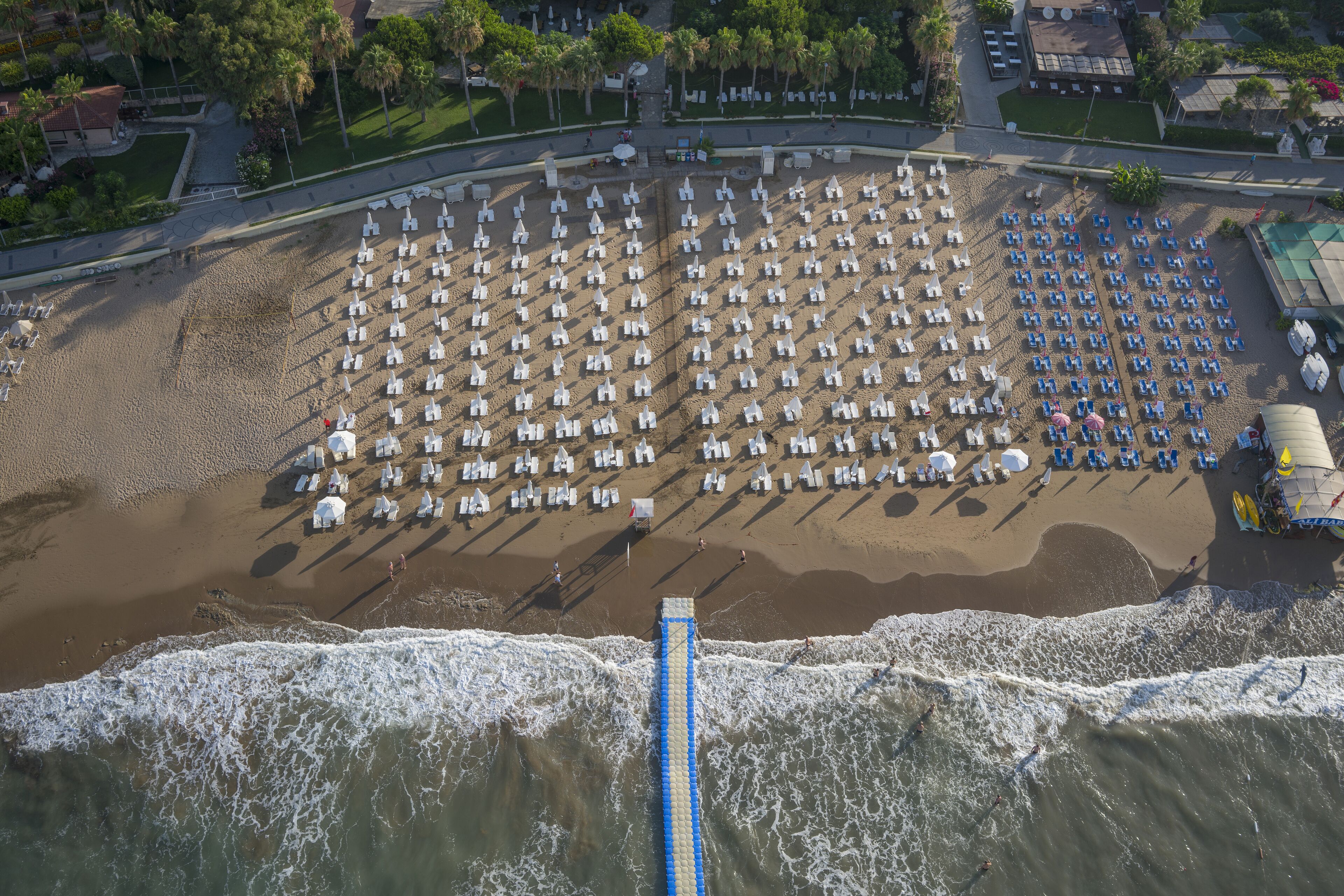 Vue aérienne d’une plage avec des rangées de chaises longues blanches et bleues, une longue promenade bleue et une zone de plage sablonneuse.