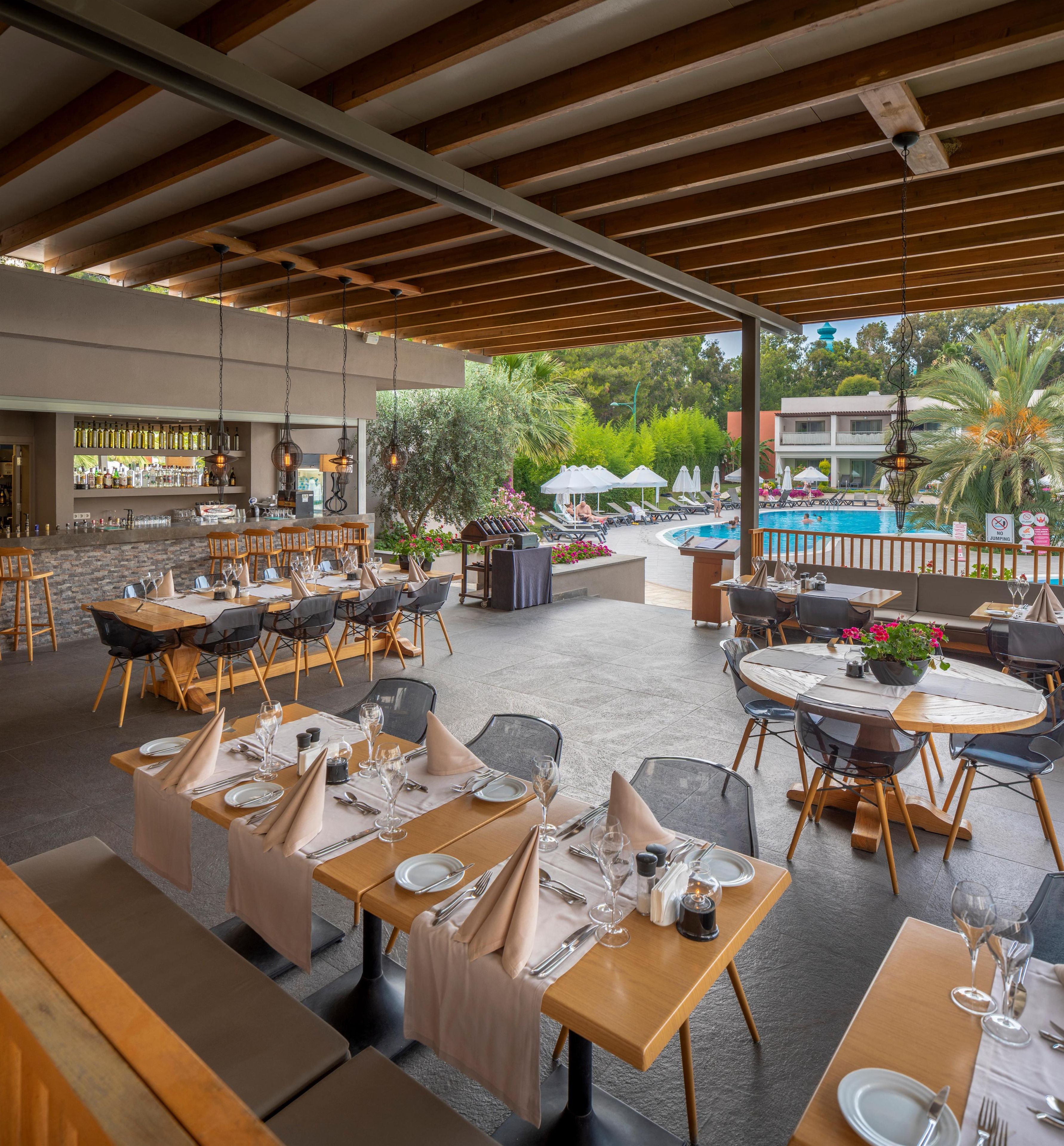 An outdoor dining area with multiple tables and chairs, a bar with a variety of bottles, and a pool in the background.