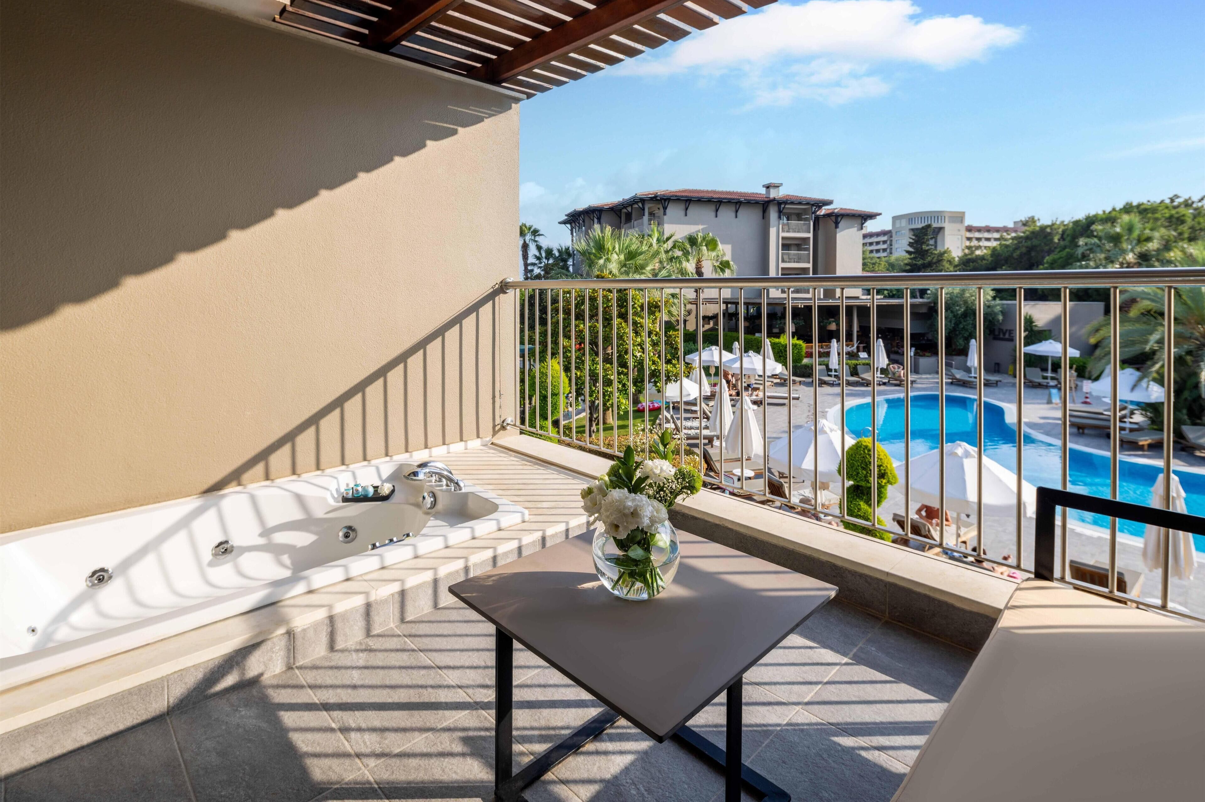 Un balcón con un jacuzzi, una mesa con un jarrón de flores y vistas a una piscina y edificios de un hotel.