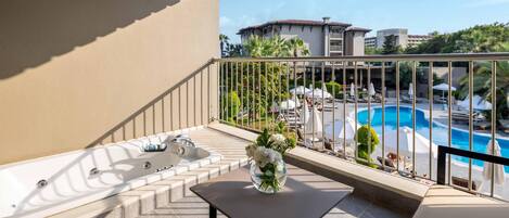 Un balcón con un jacuzzi, una mesa con un jarrón de flores y vistas a una piscina y edificios de un hotel.
