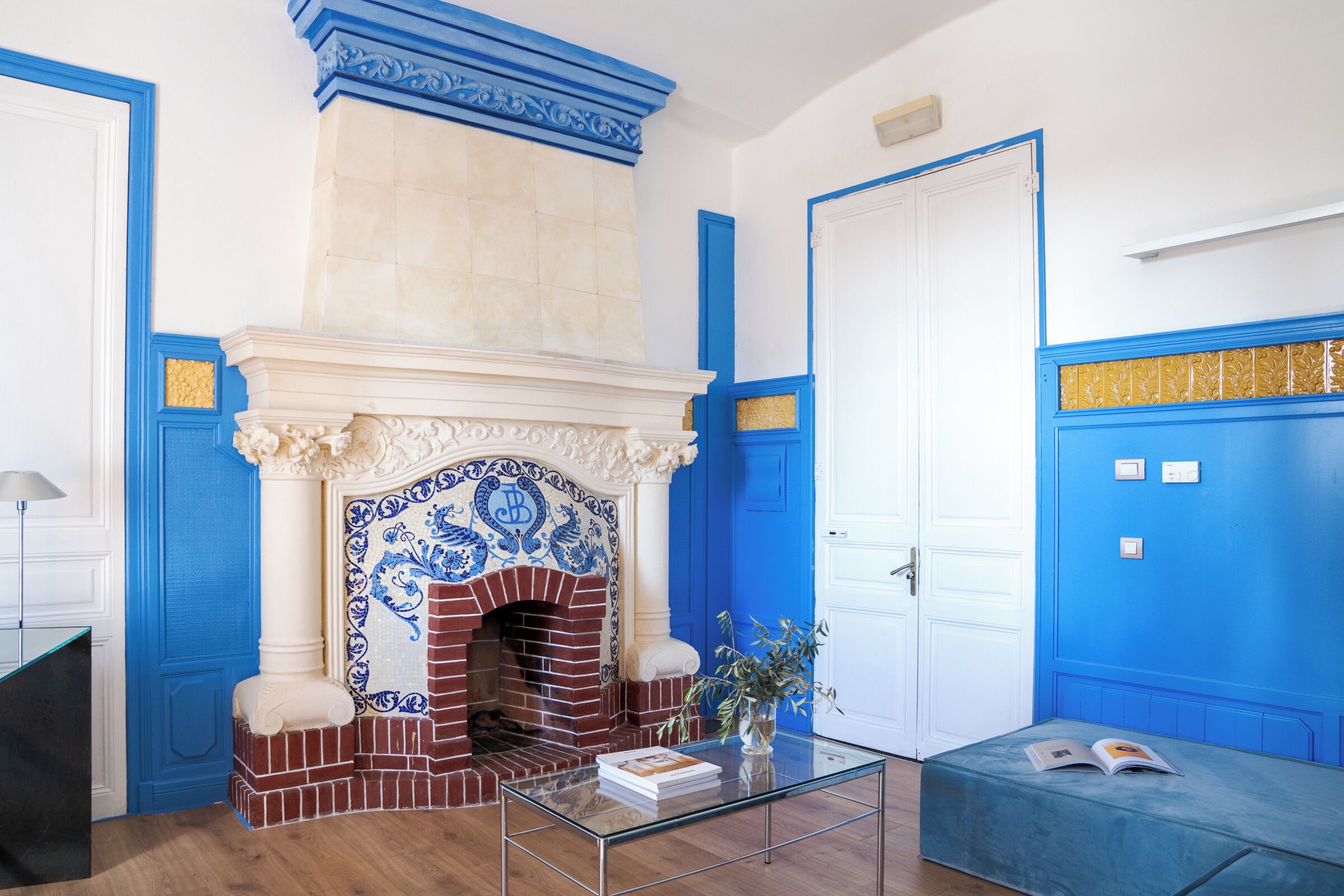 Een kamer met een open haard met blauwe en witte tegels, een witte deur en een blauwe wand met gouden sierlijsten.