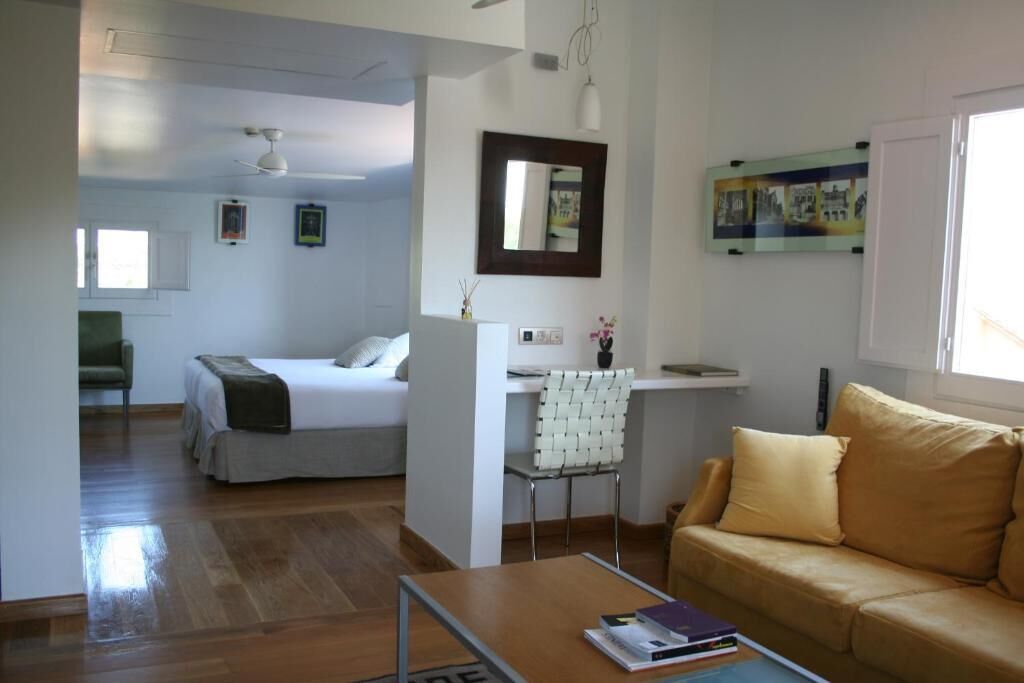 A modern, well-lit room with a bed, a sofa, a desk, and a chair. There is a window providing natural light and a wall-mounted mirror.