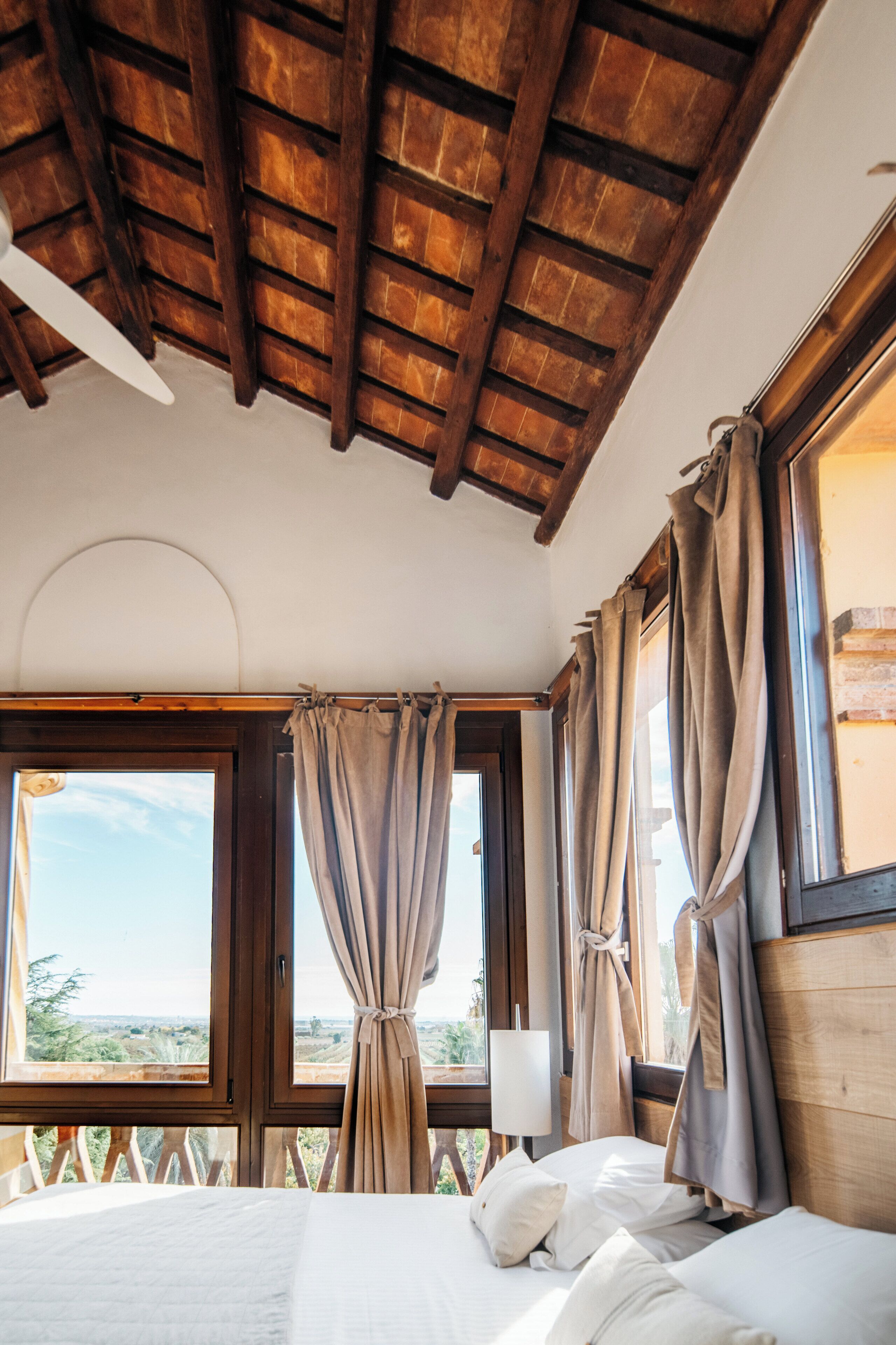 A bedroom with a wooden ceiling, large windows with curtains, a bed with white linens, and a view of the outdoors.