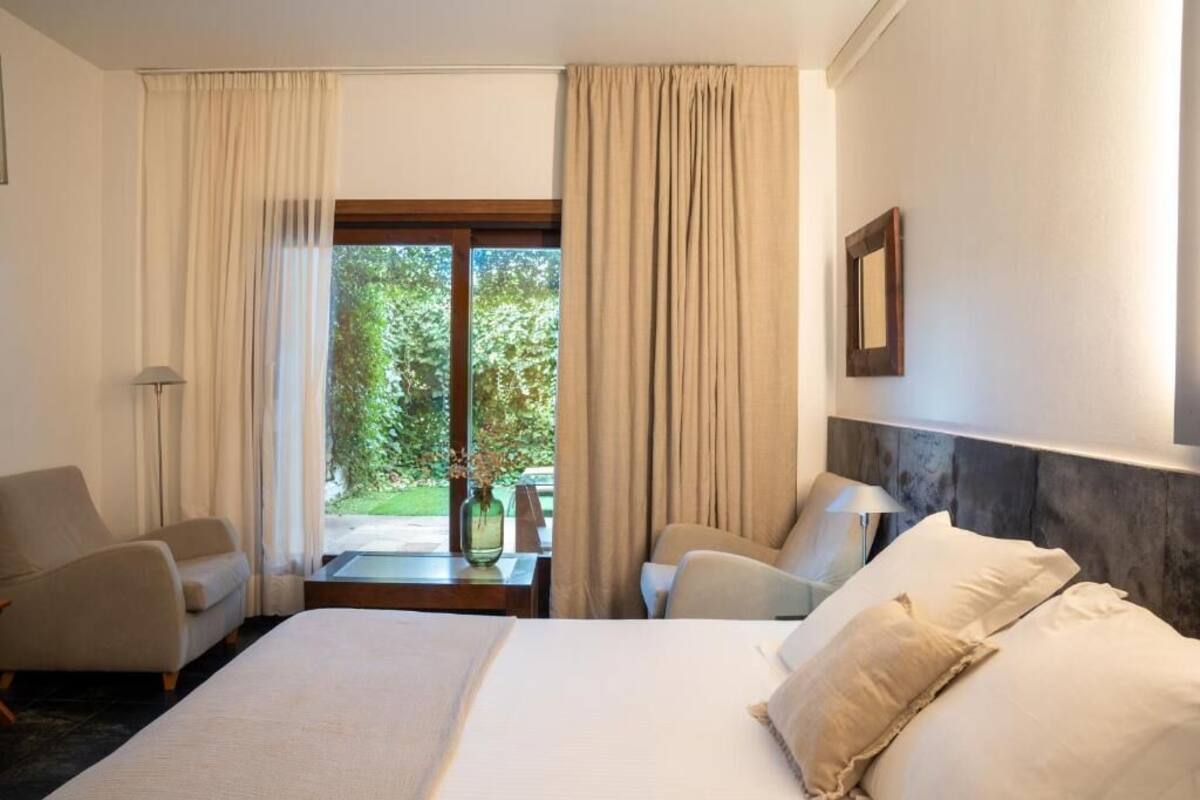 Quarto de hotel com uma cama grande, duas poltronas, uma pequena mesa e vista para o verde pela janela.