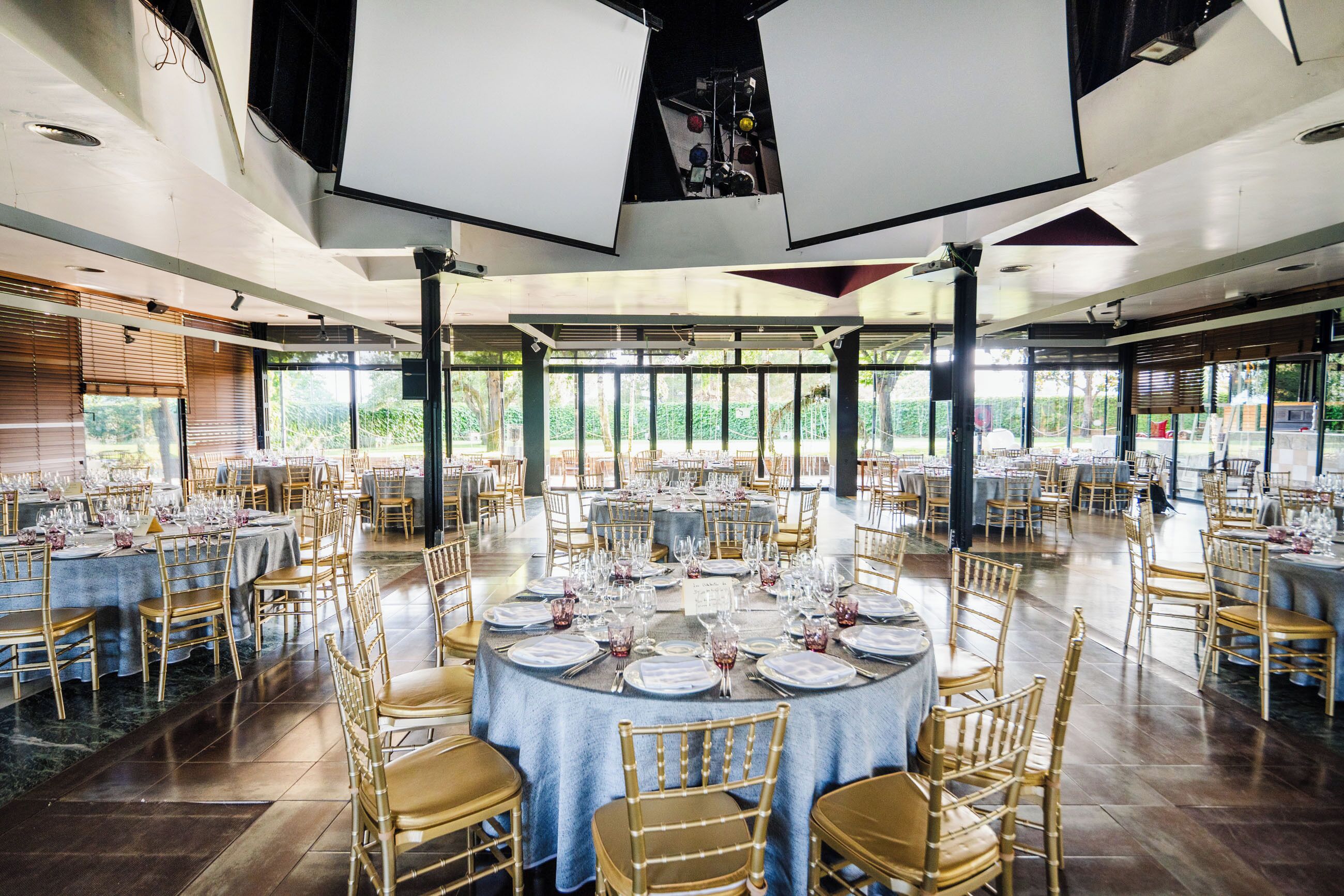 Une salle de banquet spacieuse, dotée de nombreuses tables rondes dressées pour le repas, de grandes fenêtres laissant entrer la lumière naturelle et d’un plafond haut équipé de luminaires.