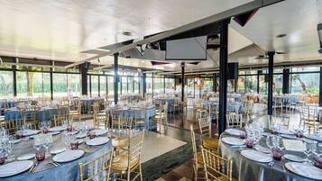 Un amplio salón de banquetes con varias mesas redondas dispuestas para cenar, ventanas grandes que ofrecen vistas a zonas verdes y un techo alto con luminarias de estilo industrial.
