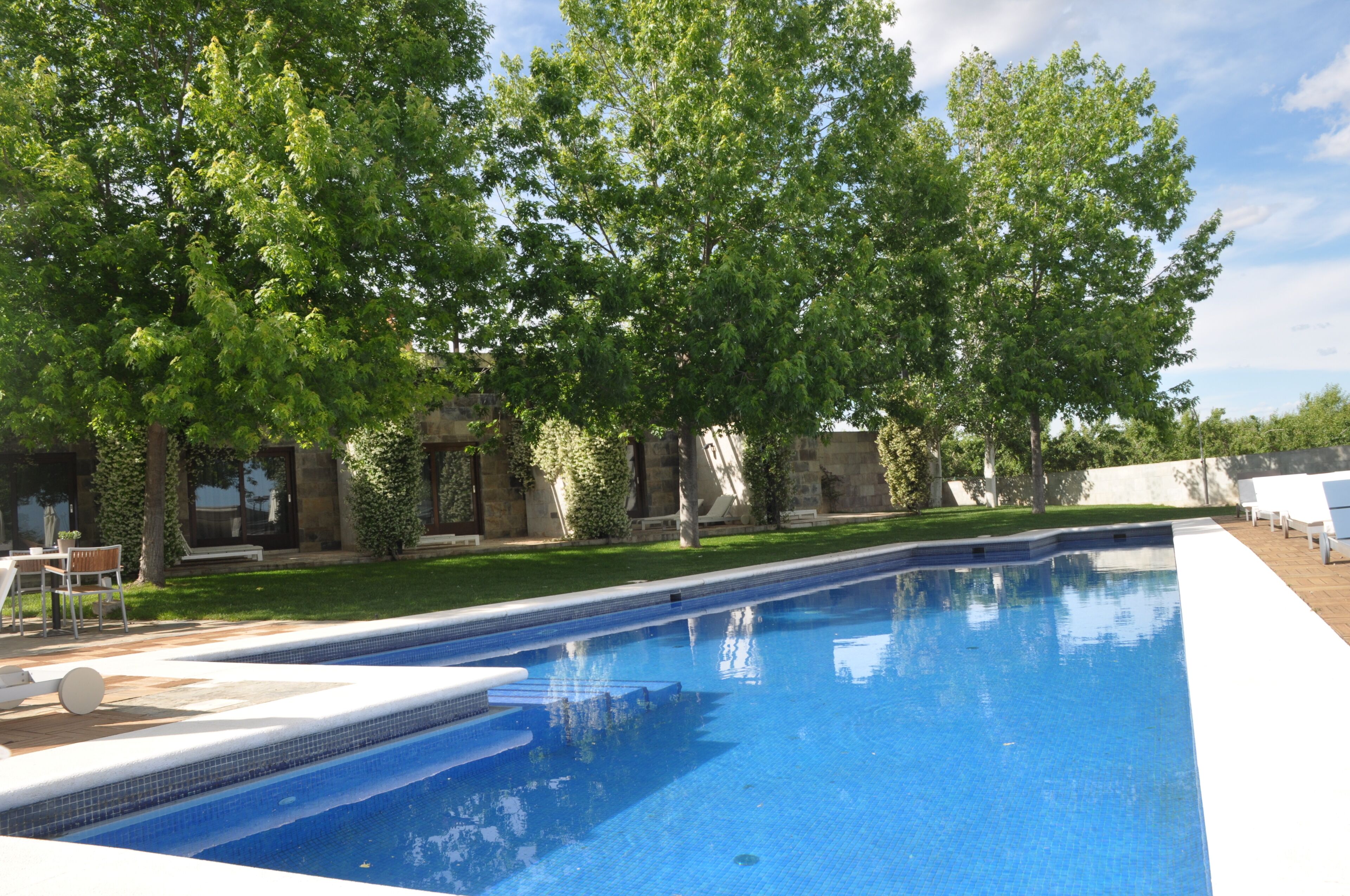 Una piscina con borde de baldosas, rodeada de árboles y un edificio de piedra.