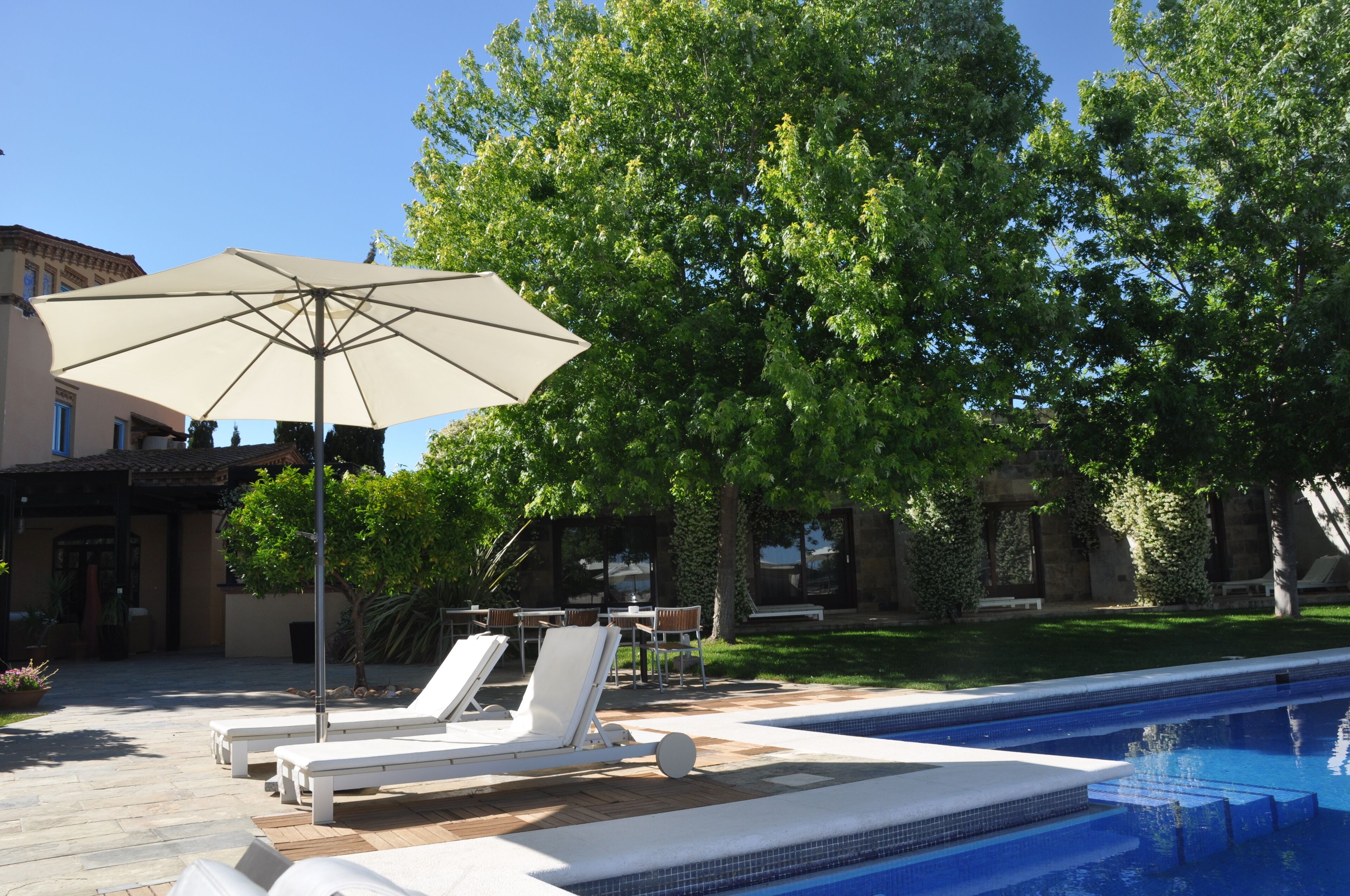 Zona junto a la piscina con sillas de descanso y una sombrilla grande, rodeada de exuberante vegetación y un edificio con tejado de baldosas.