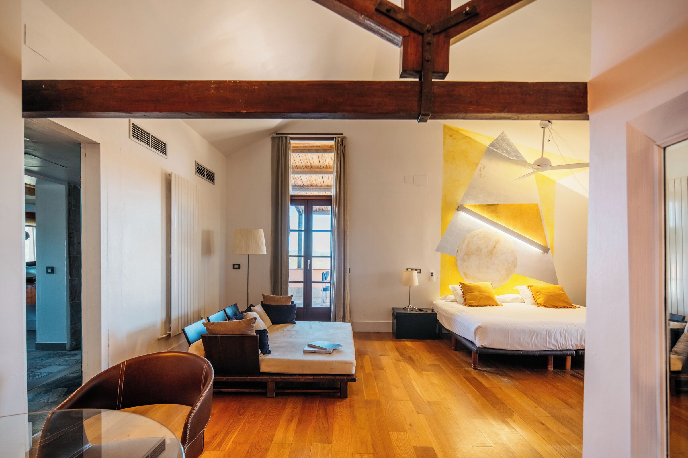 A spacious room with a wooden floor, a bed with a white duvet, a sofa with cushions, and a small table.