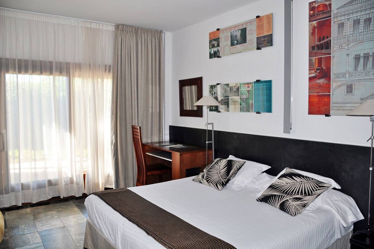 Quarto de hotel com cama, escrivaninha, cadeira e janela com cortinas.