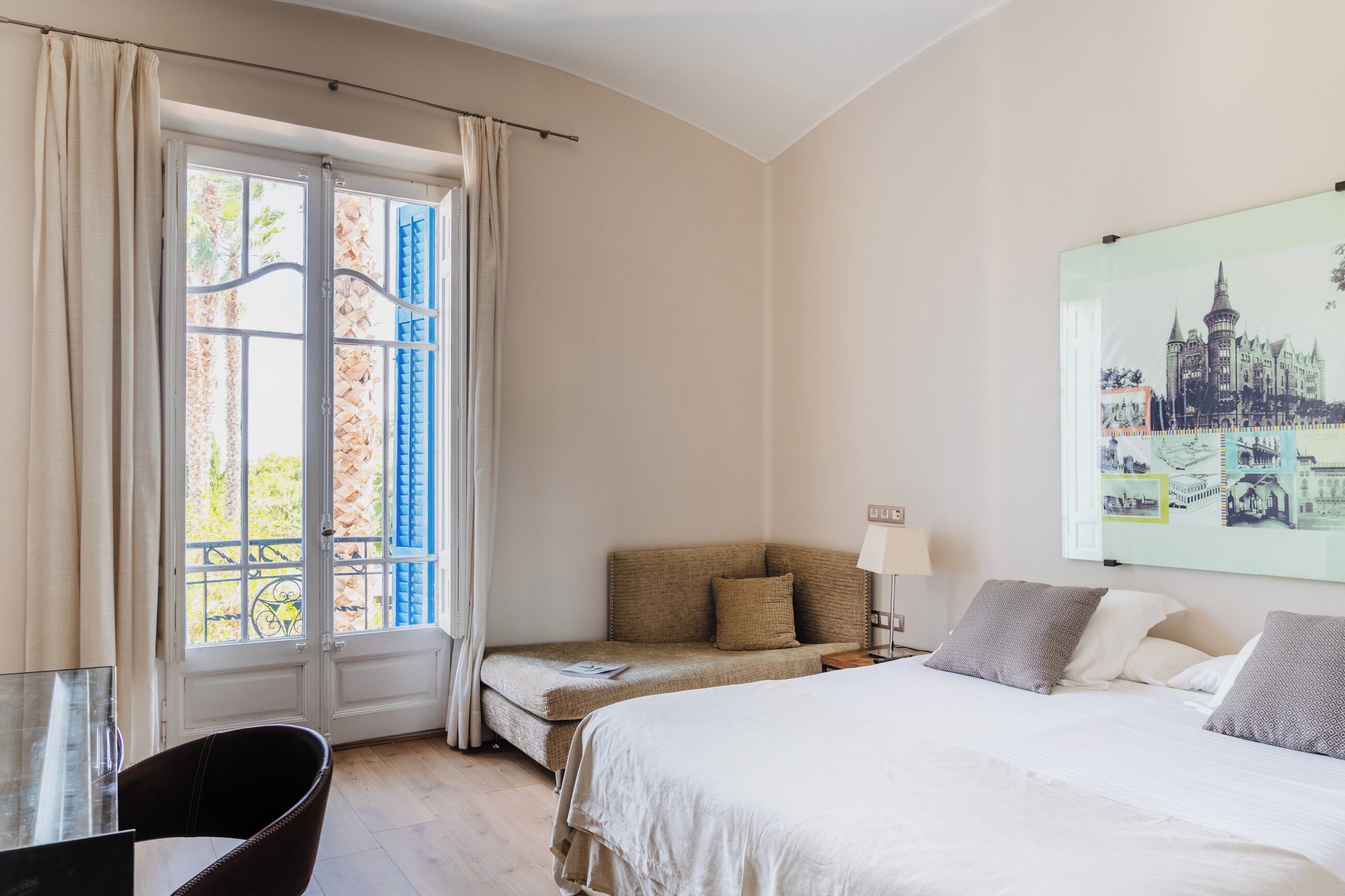 A hotel room with a bed, a small seating area, a window with curtains, and a wall-mounted picture.