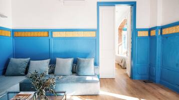 Una habitación con decoración azul y blanca, un sofá con cojines, una mesa de centro de cristal y una puerta que da a otra habitación.