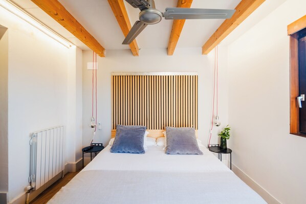 A bedroom with a bed, wooden ceiling beams, a ceiling fan, a radiator, and a bedside table with a plant.