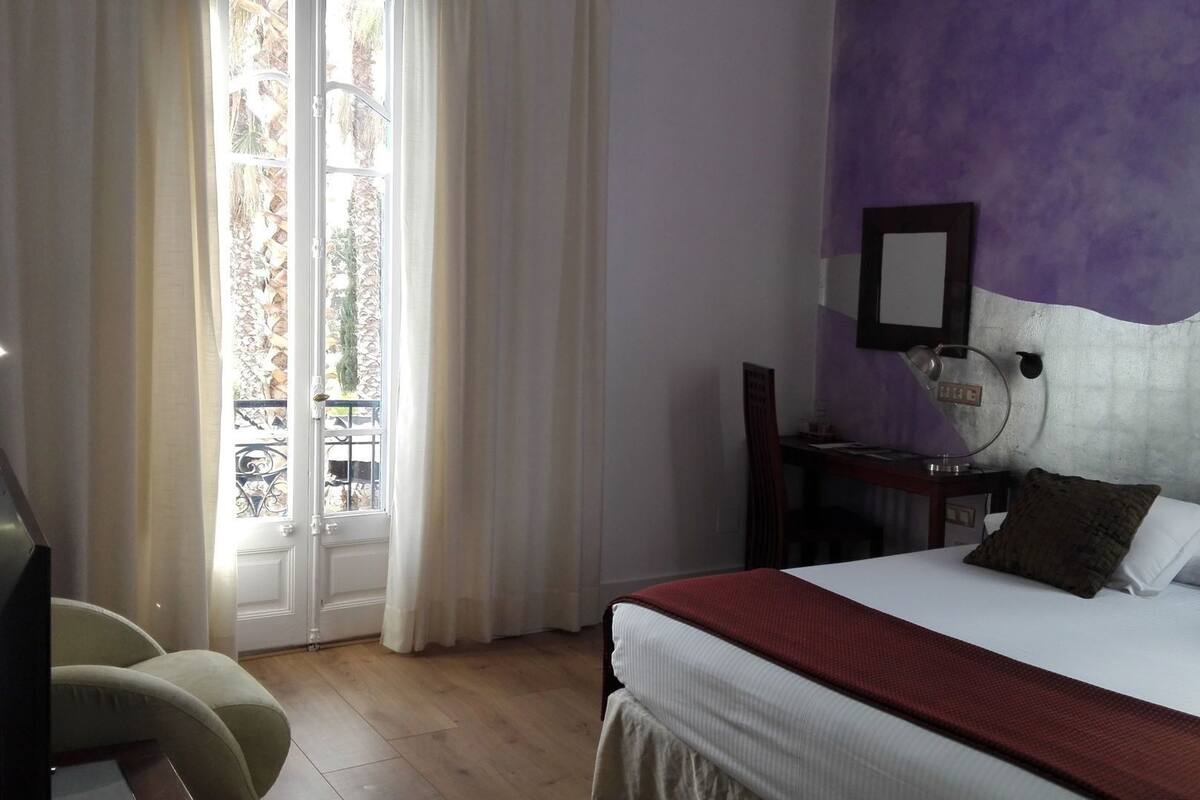 Quarto de hotel com cama, escrivaninha com luminária, cadeira e janela com cortinas.
