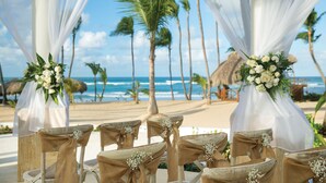 Decoració de boda a la platja amb cadires blanques i teixits penjats, palmeres i l'oceà al fons.